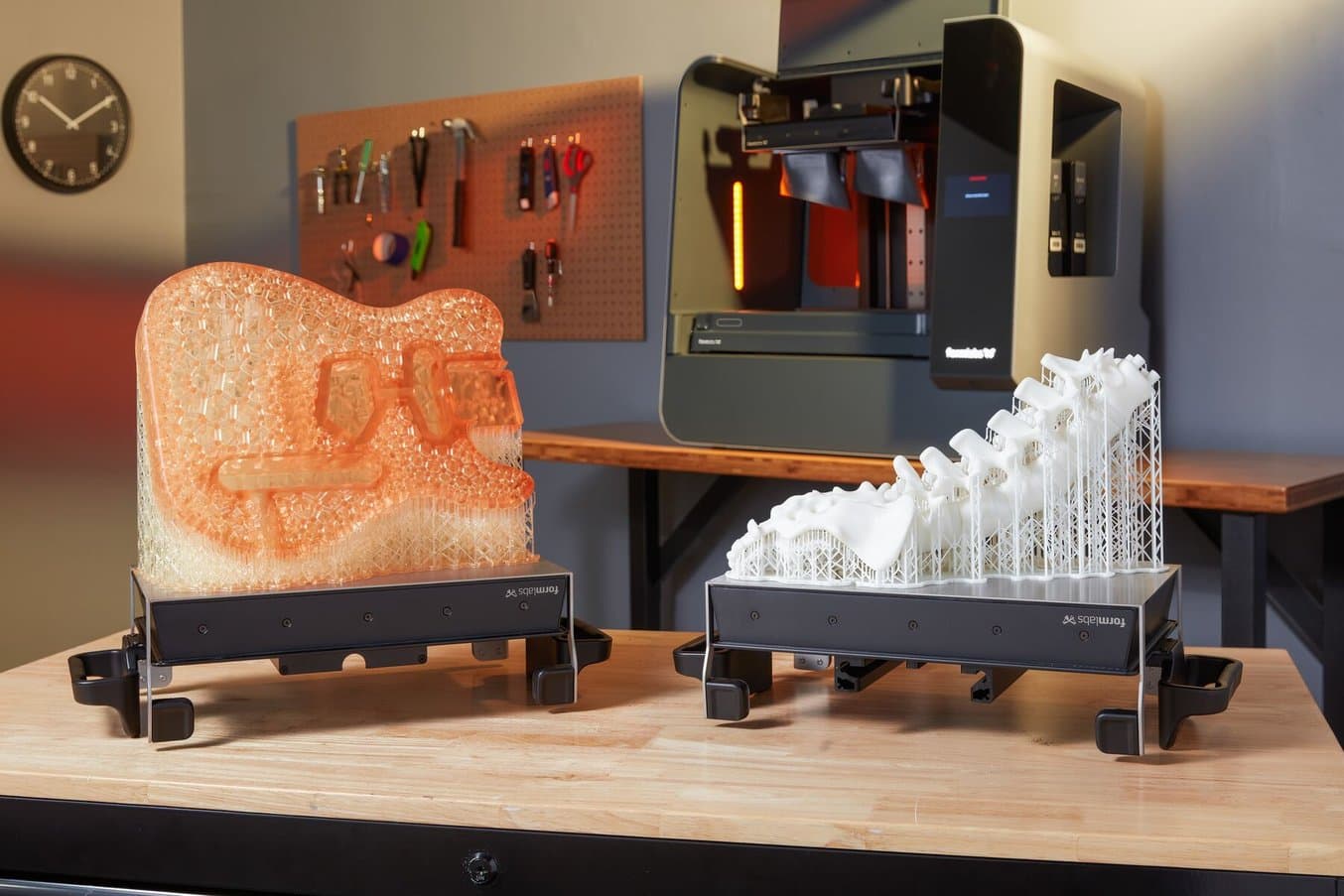 A 3D printed guitar frame and a 3D printed anatomical model of a spine on build platforms sit on a table in front of a Form 3L stereolithography (SLA) 3D printer.