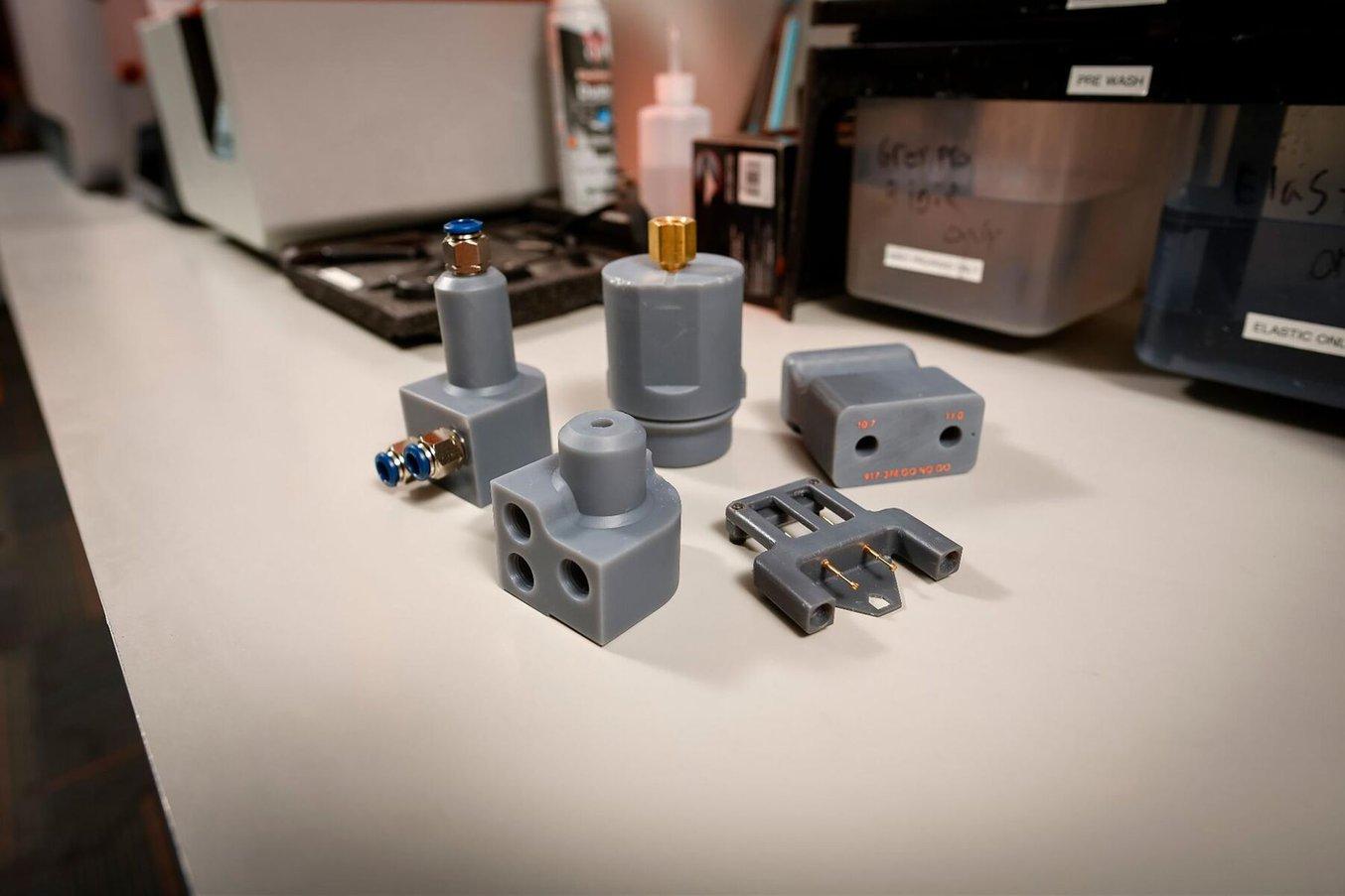 Five small jigs, some with off-the-shelf hardware incorporated, sit on a table in front of a 3D printing post-processing station.