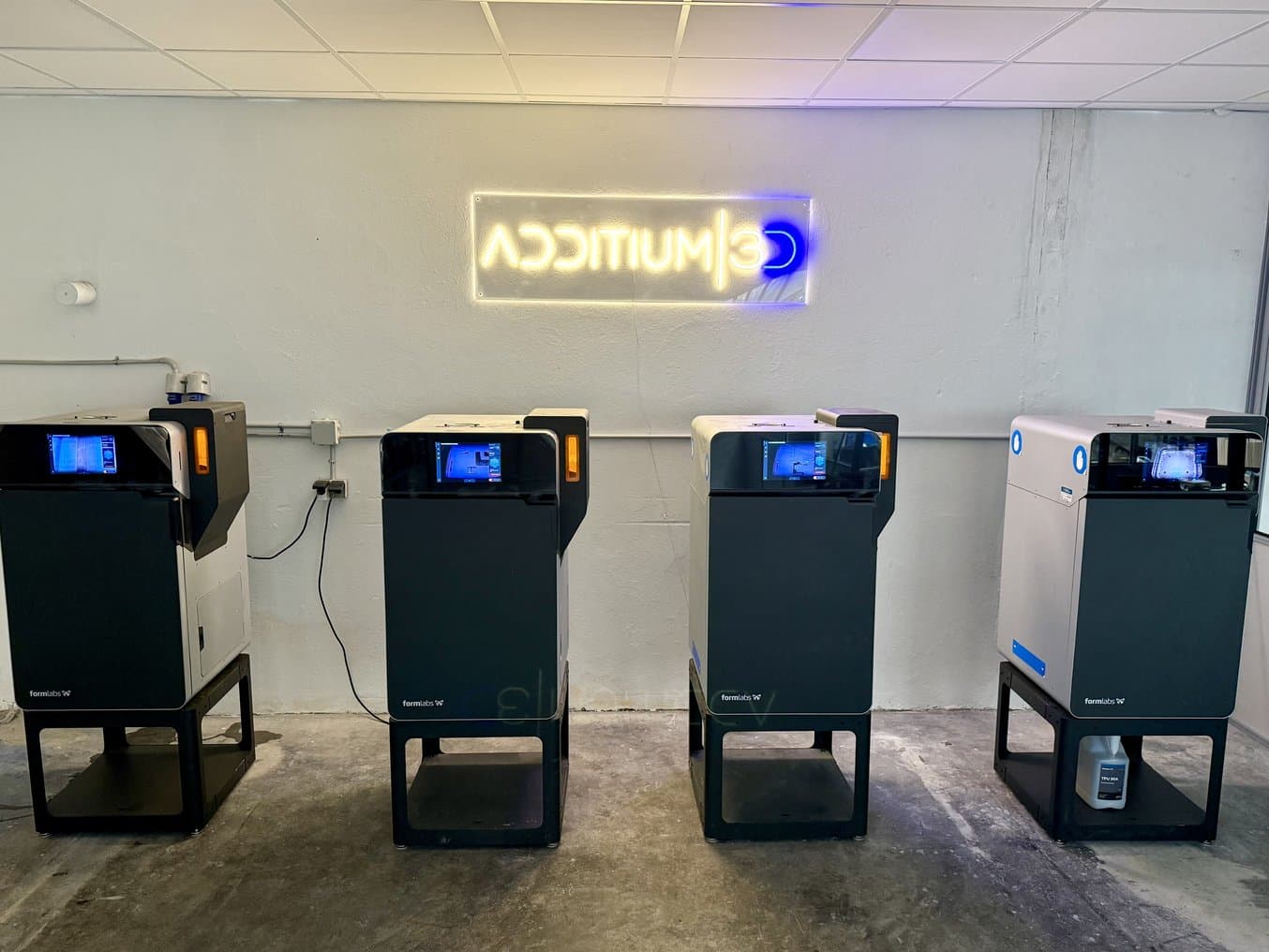 四台 Fuse 1+ 30W SLS 3D 打印机位于一个写着“Additium3D”的霓虹灯下
